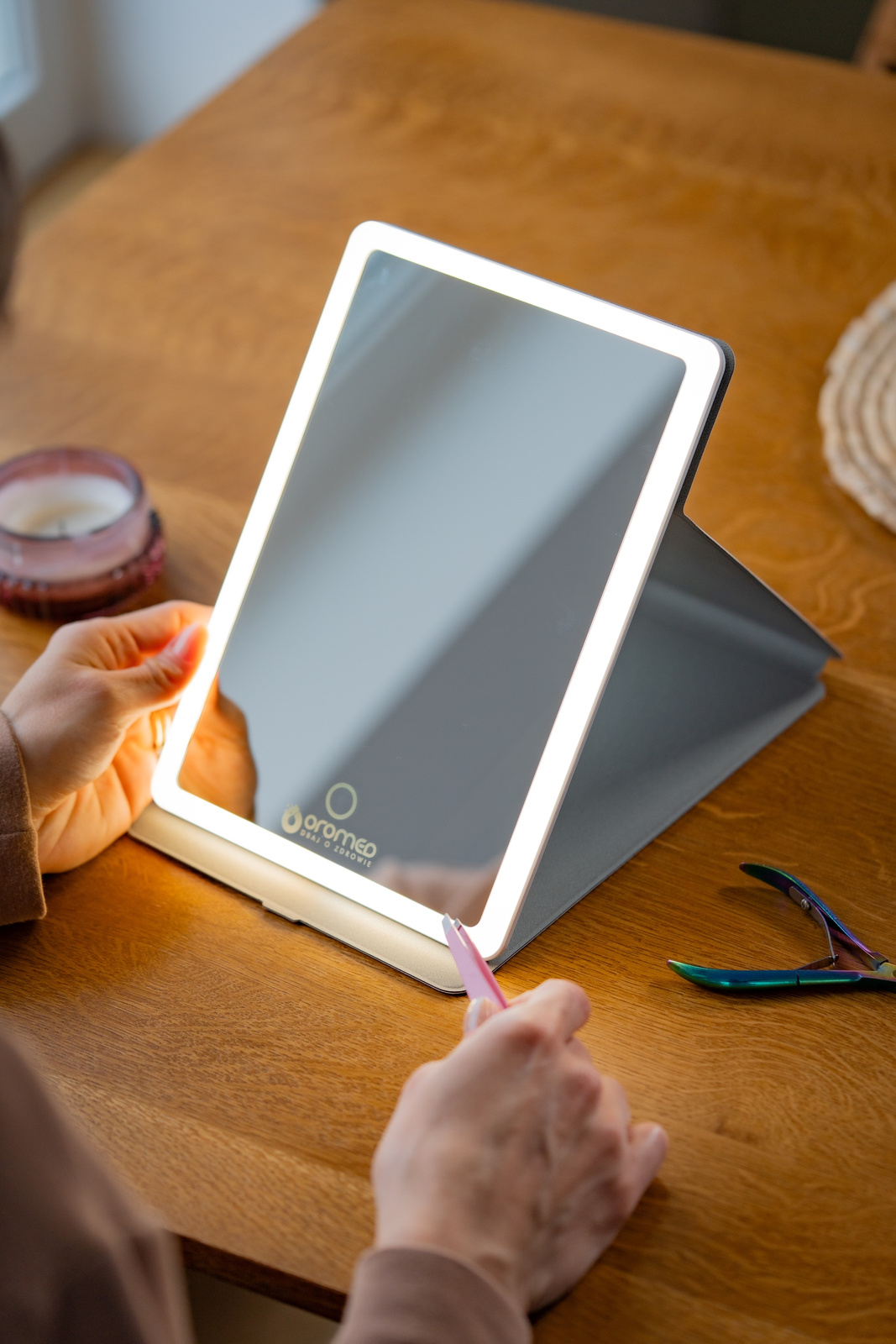 Podświetlane lustro z osobą trzymającą pęsetę, druga ręka trzyma różowe narzędzie. Świeca i cążki do paznokci na drewnianym stole.