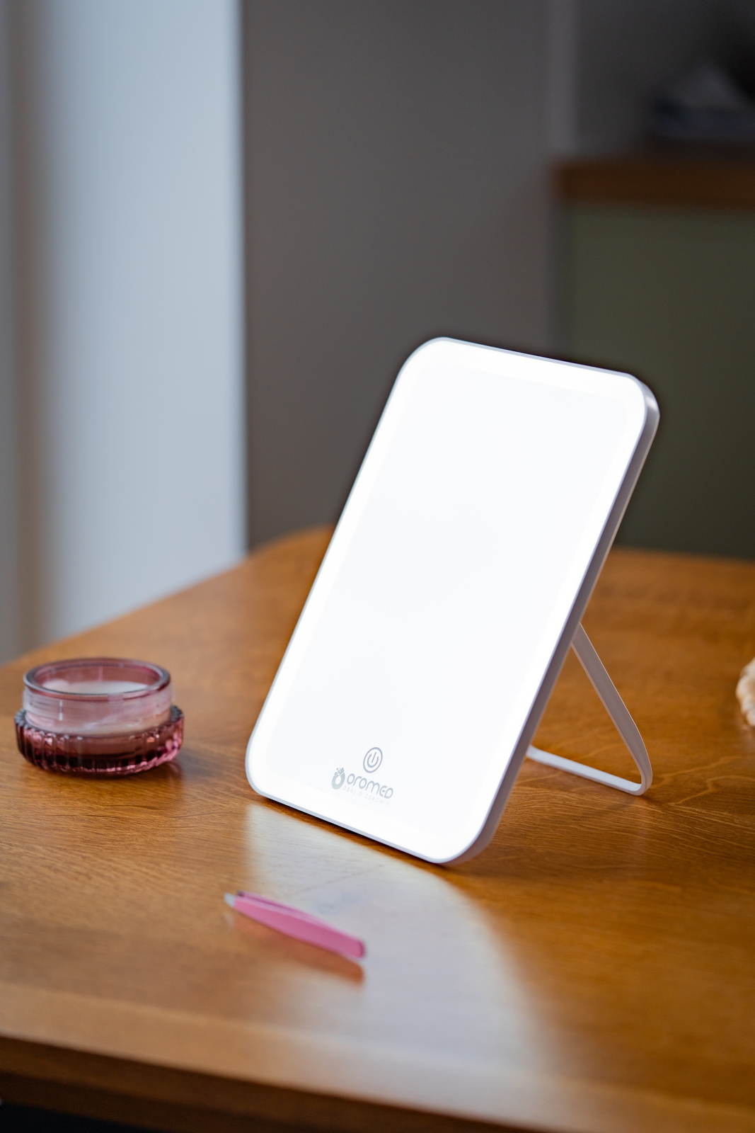 Podświetlane białe lustro stoi na drewnianym stole. Na stole słoik i pęseta. Tło jest rozmyte.