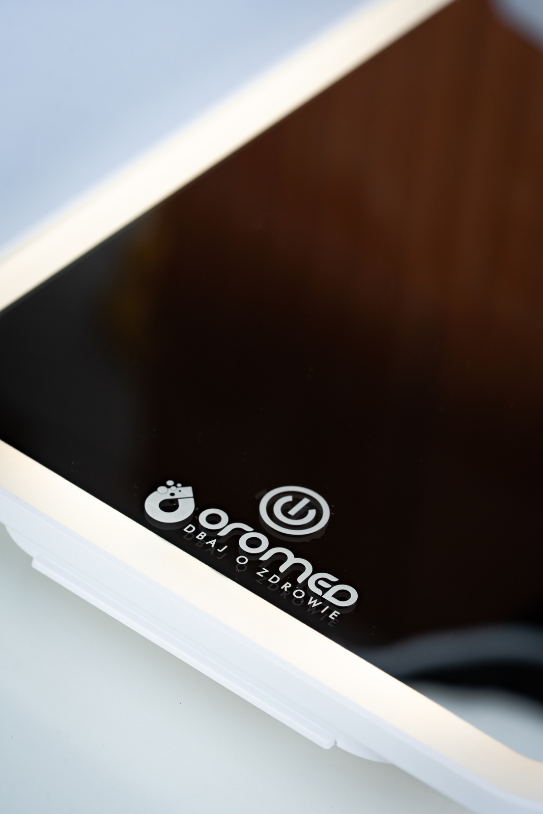 Zbliżenie białego urządzenia z czarnym ekranem. Urządzenie ma logo 'oromed DBAJ O ZDROWIE' i przycisk zasilania.