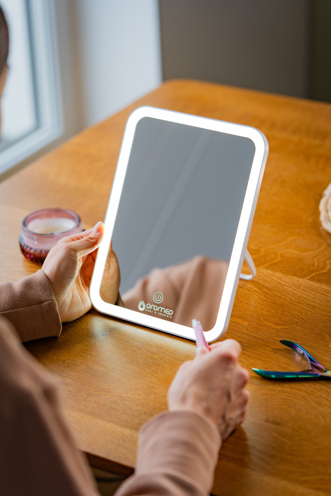 Osoba trzyma podświetlane lustro na drewnianym stole. Na stole leżą różowy długopis i cążki do paznokci. Świeca znajduje się w tle.