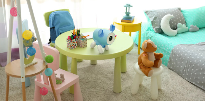 Pokój dziecięcy ze stołem, krzesłami, łóżkiem i zabawkami. Na stole stoi niebieski projektor. Miś siedzi na stołku.