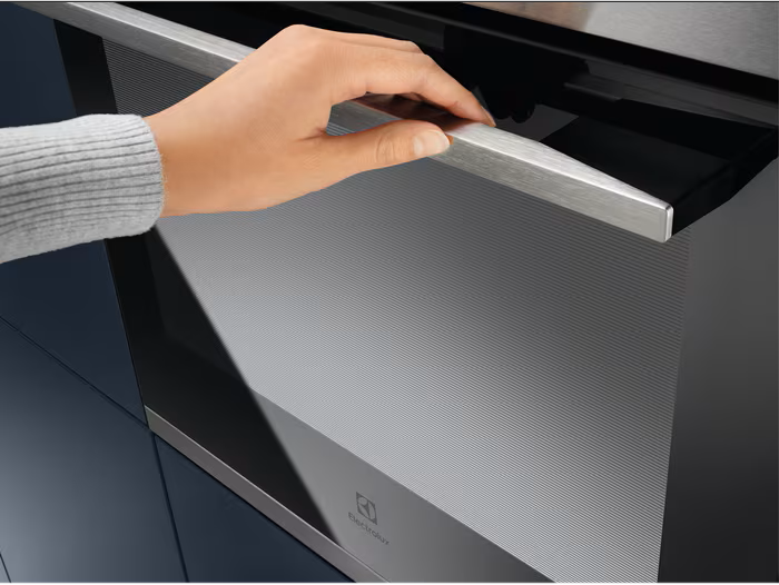 Eine Hand öffnet eine Backofentür. Der Ofen ist schwarz und Edelstahl. Der Schrank ist dunkelblau. Die Marke ist Electrolux.