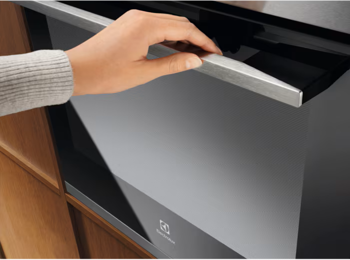 Eine Hand öffnet eine schwarze Ofentür mit einem silbernen Griff. Holzschrank links. Electrolux-Logo sichtbar.