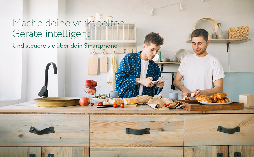Zwei Männer in einer Küche bereiten Essen zu. Einer benutzt ein Telefon. Croissants, Brot, Früchte auf der Theke.