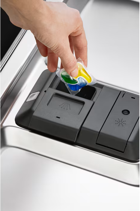 Eine Hand legt einen mehrfarbigen Geschirrspülmittel-Pod in den Spender einer Spülmaschine. Der Spender ist dunkelgrau.