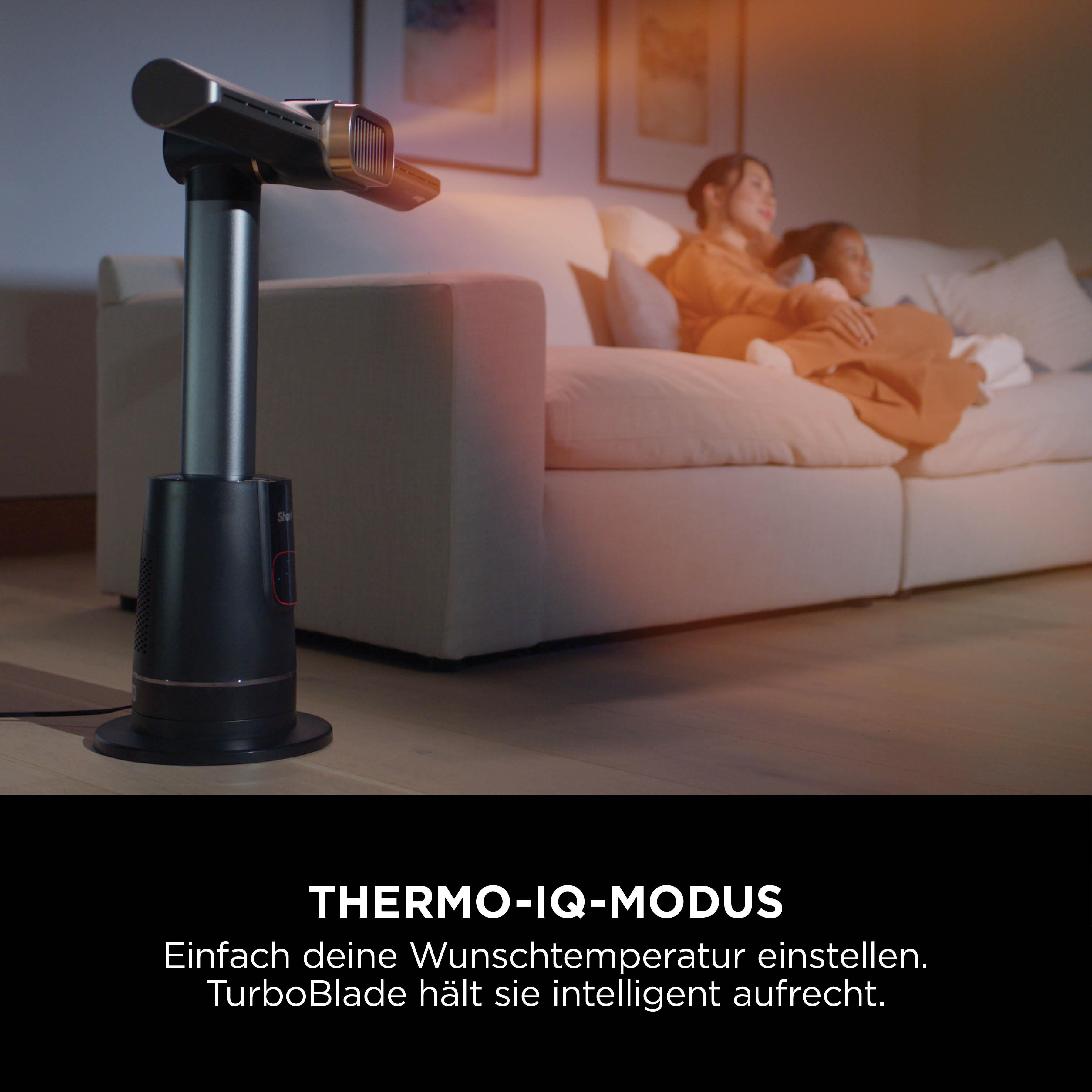 Ein schwarzer und silberner Ventilator steht in einem Raum. Zwei Personen sitzen auf einem weißen Sofa. Der Text lautet Thermo-IQ-Modus.