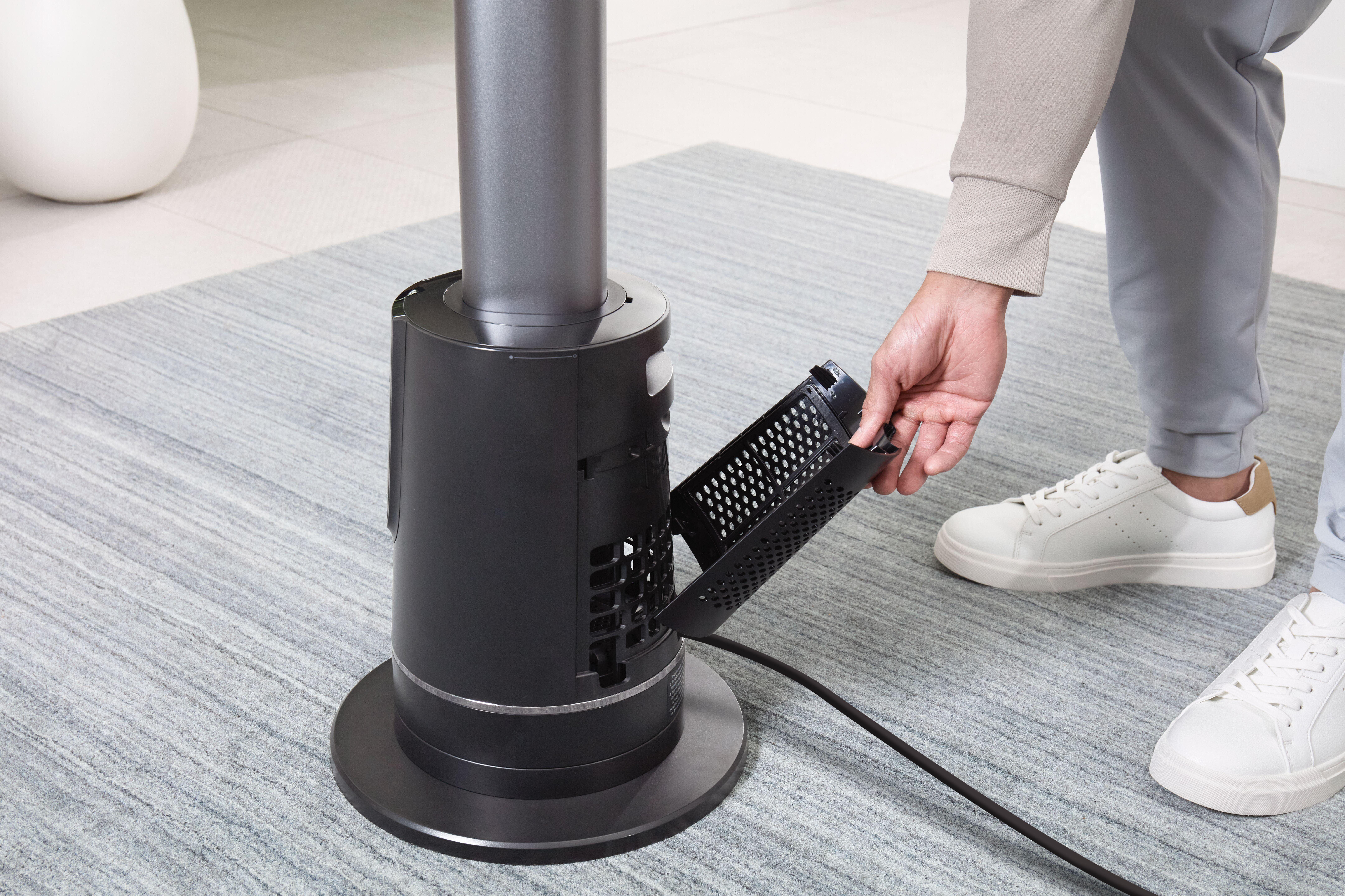 Eine Person entfernt einen Filter von einem schwarzen Ventilator. Sie trägt graue Hosen und weiße Schuhe. Der Ventilator steht auf einem grauen Teppich.