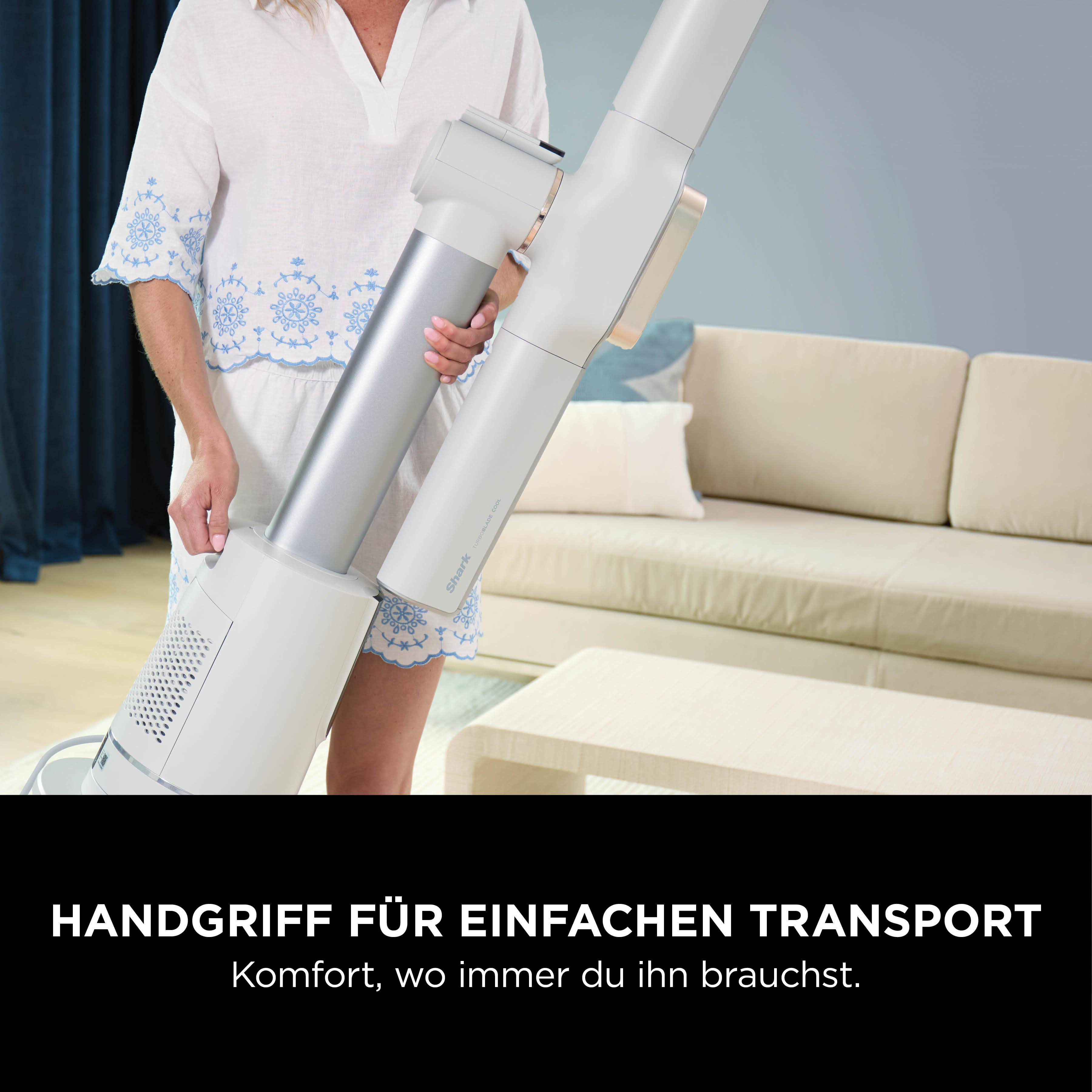 Eine Frau hält einen weißen und silbernen Staubsauger. Sie befindet sich in einem Raum mit einer Couch und einem blauen Vorhang.