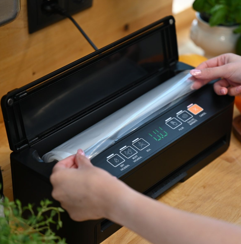 Osoba wkłada rolkę plastiku do czarnej zgrzewarki próżniowej do żywności. Urządzenie znajduje się na drewnianej powierzchni z zielonym wyświetlaczem.