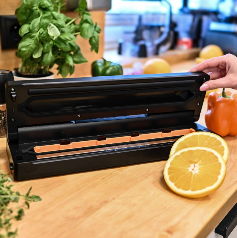 Scena kuchenna z zgrzewarką próżniową, plasterkami pomarańczy i warzywami. Ręka otwiera zgrzewarkę. Widoczna bazylia i papryka.