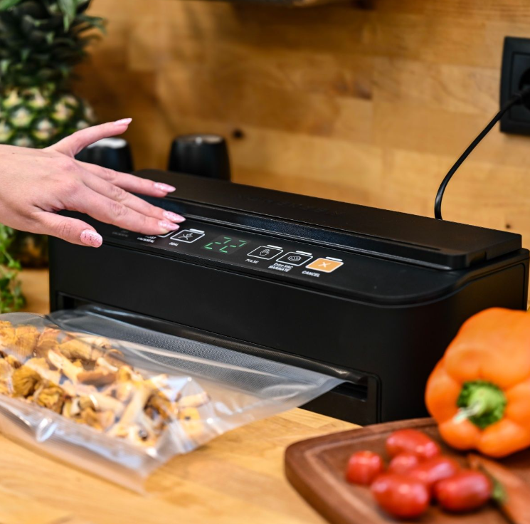 Dłoń naciska przycisk na czarnej zgrzewarce próżniowej. W środku jest worek z jedzeniem. W pobliżu znajduje się papryka i pomidory. Tło jest drewniane.