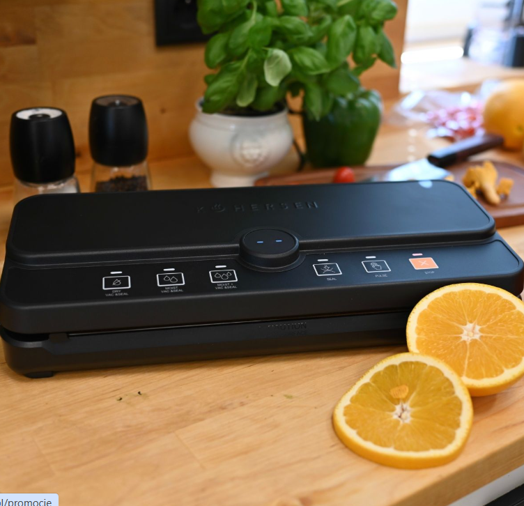 Czarna zgrzewarka próżniowa z plasterkami pomarańczy na drewnianej powierzchni. Zioła i inne przedmioty kuchenne w tle.