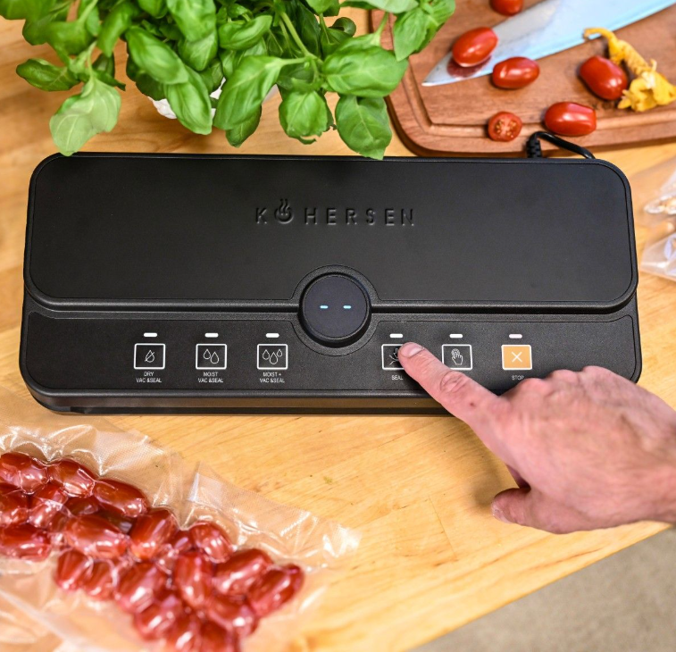 Czarna zgrzewarka próżniowa z ręką naciskającą przycisk zgrzewania. Pomidory, bazylia i deska do krojenia w tle.