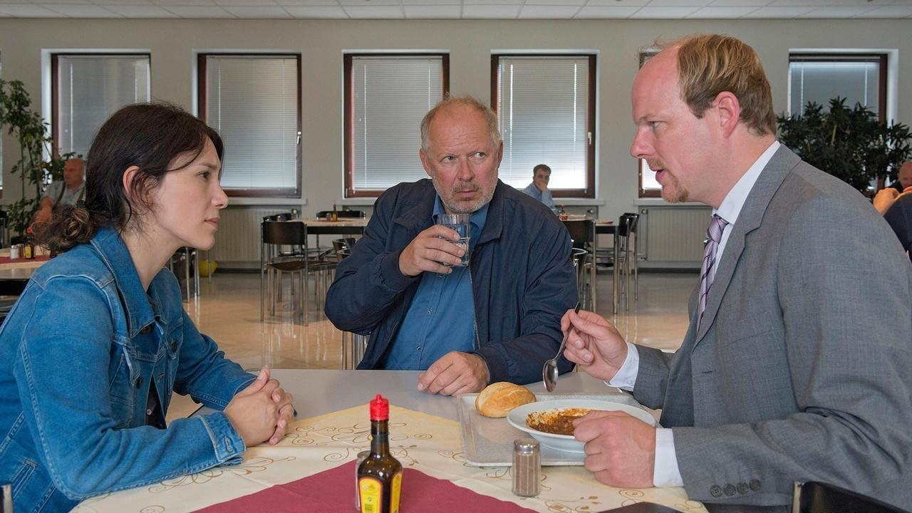 Drei Personen sitzen in einer Cafeteria an einem Tisch. Eine Frau, zwei Männer. Essen und Getränke sind vorhanden. Fenster im Hintergrund.