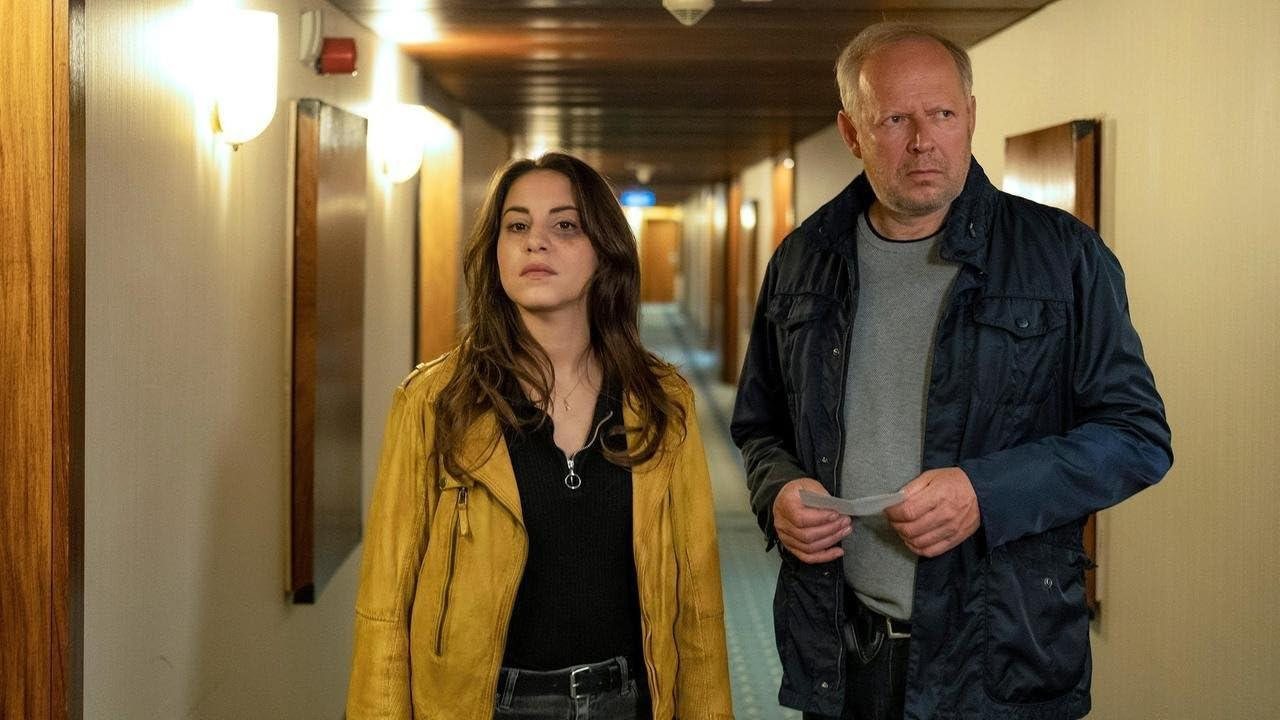 Eine Frau in einer gelben Jacke und ein Mann in einer blauen Jacke gehen einen Flur entlang. Sie befinden sich in einem schwach beleuchteten Raum.