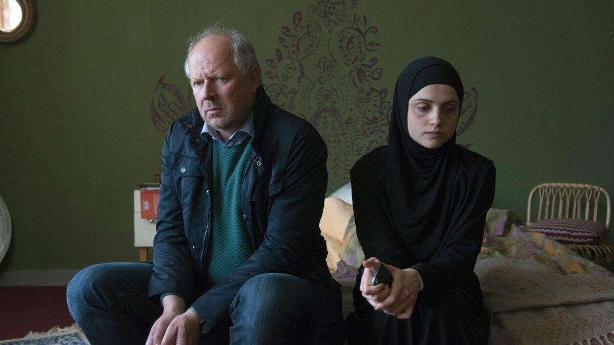 Ein Mann und eine Frau mit schwarzem Hidschab sitzen drinnen. Der Mann trägt eine blaue Jacke und einen Pullover. Hinter ihnen ist eine grüne Wand.