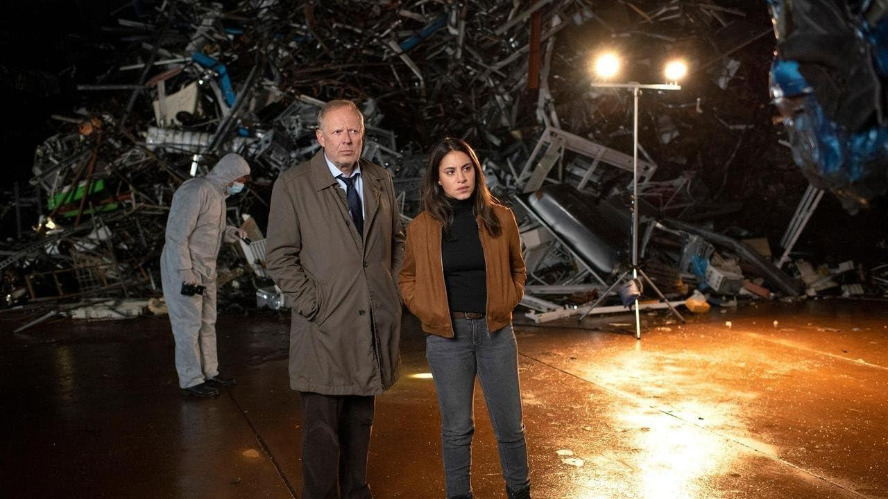 Zwei Personen stehen vor einem Metalltrümmerhaufen. Eine Person in einem Schutzanzug untersucht die Szene. Ein Licht beleuchtet den Bereich.
