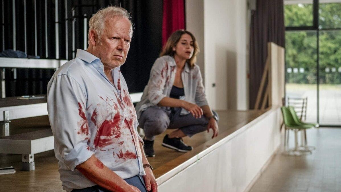 Ein älterer Mann mit Blut auf dem Hemd wirkt besorgt. Eine Frau hockt hinter ihm, ebenfalls mit Blut. Im Hintergrund ist eine Bühne zu sehen.
