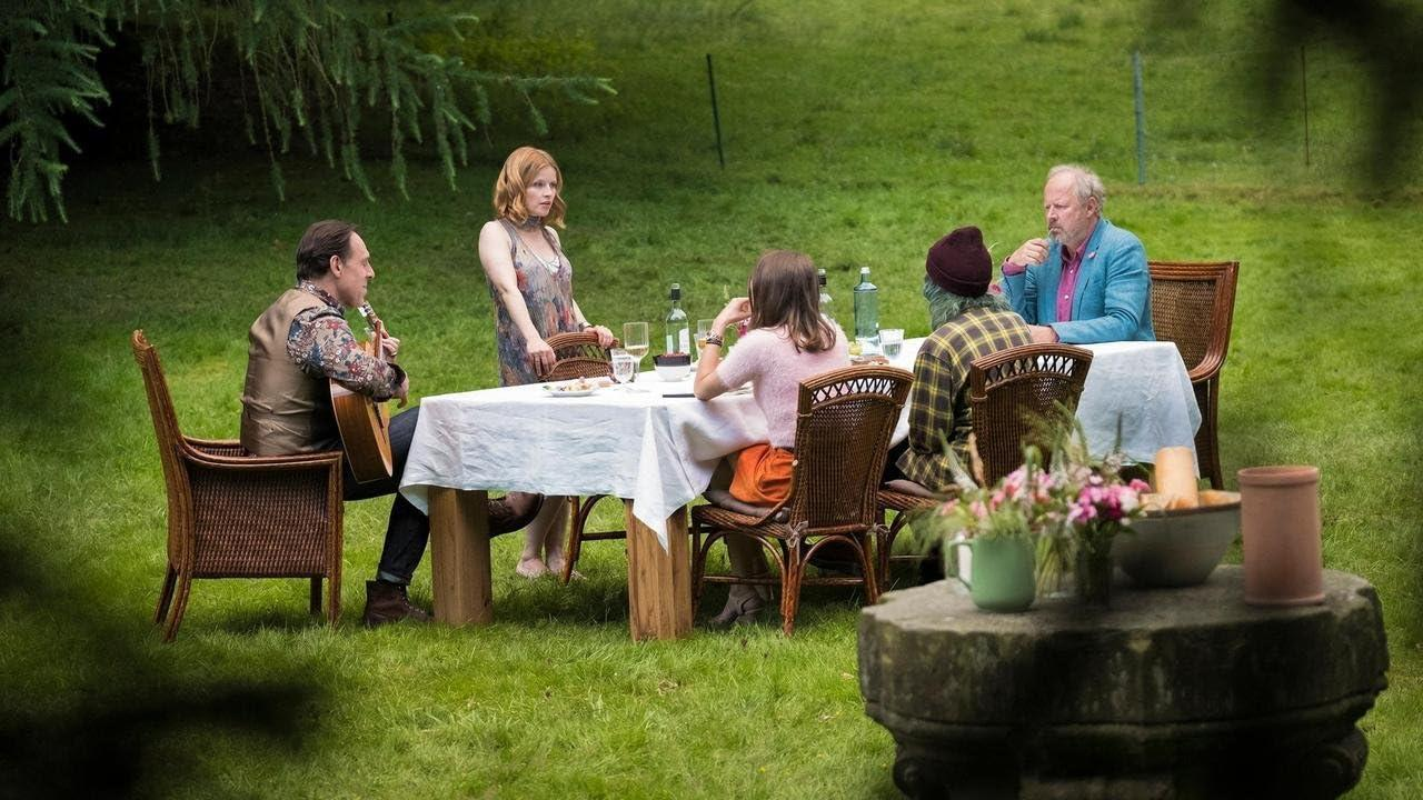 Menschen sitzen in einem Garten an einem Tisch. Ein Mann spielt Gitarre. Auf dem Tisch stehen Essen, Getränke und Blumen.