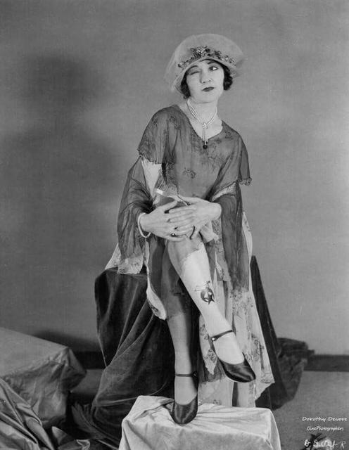 Eine Frau in einem Flapper-Kleid und Hut zwinkert, sitzt mit übereinandergeschlagenen Beinen. Sie hat ein Tattoo auf ihrem Bein und schwarze Schuhe, in Schwarzweiß.