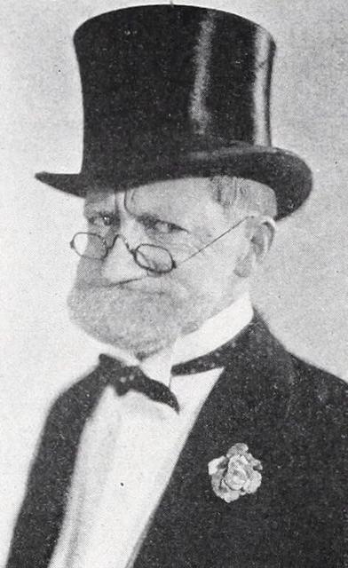 Ein älterer Mann mit Zylinder, Brille und Anzug. Er hat einen weißen Bart, eine Fliege und eine Blume am Revers.