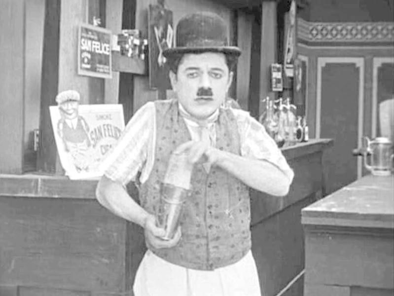 Ein Mann mit Schnurrbart und Melone hält einen Cocktailshaker. Er trägt eine Weste und ein weißes Hemd, hinter einer Bar.