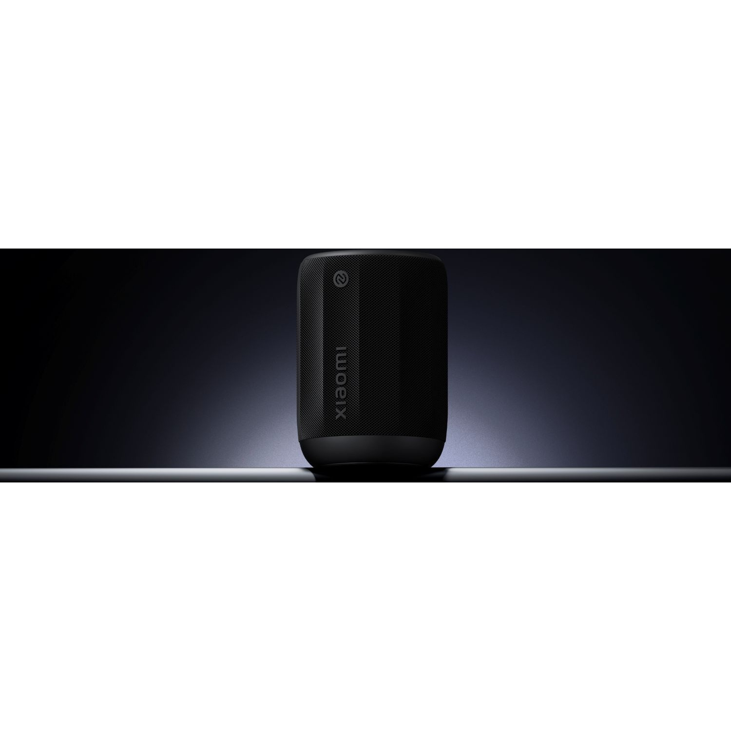 A black Xiaomi speaker stands on a reflective surface against a dark gradient background. The speaker is cylindrical.