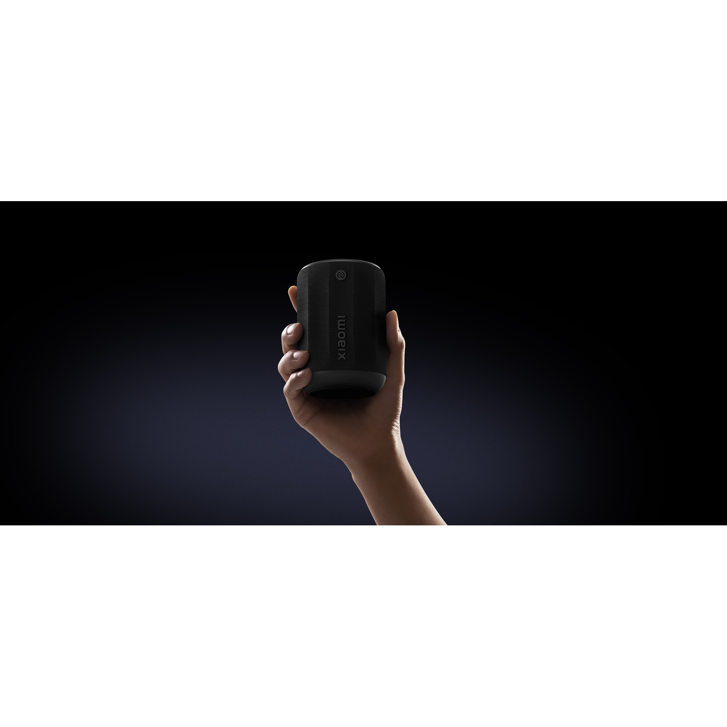 A hand holds a black Xiaomi speaker against a dark blue background. The speaker is cylindrical with Xiaomi branding on it.