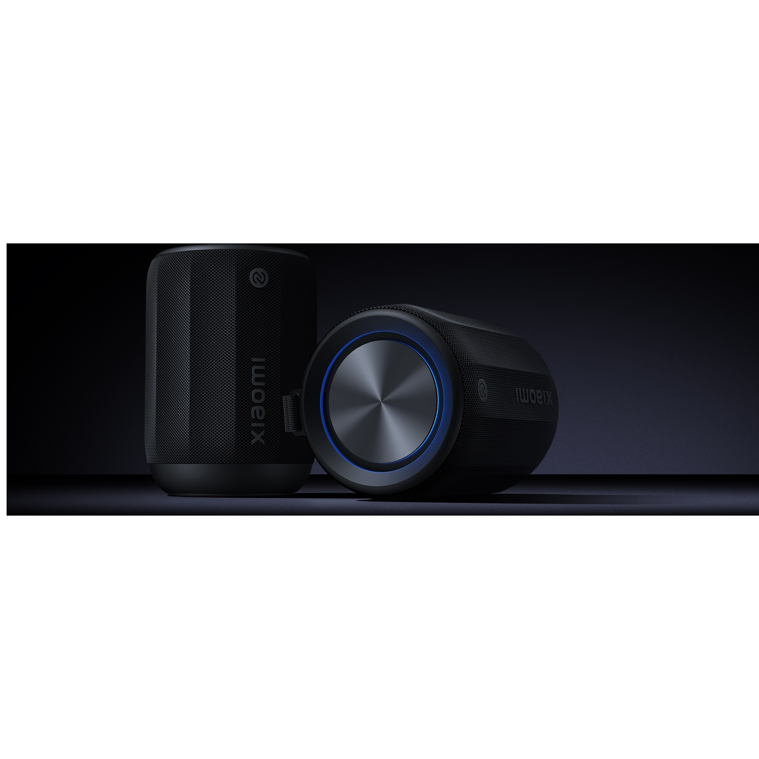 Two black Xiaomi speakers. One upright, one on its side. Blue light accents the side speaker. The background is a dark gradient.