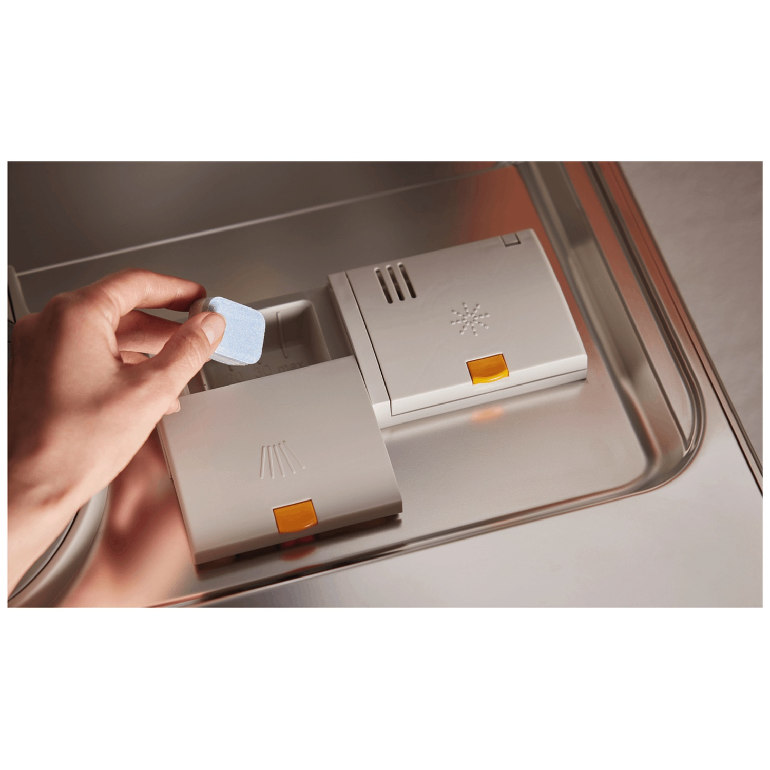 A hand places a blue dishwasher tablet into the dispenser. Two white compartments with orange buttons sit in a stainless steel machine.
