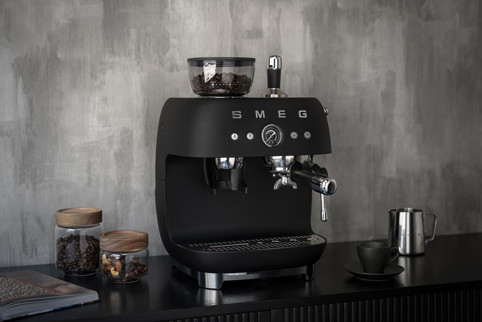 Schwarze SMEG Espressomaschine mit Kaffeebohnen, Tassen und einem Edelstahlkrug auf einer schwarzen Oberfläche.