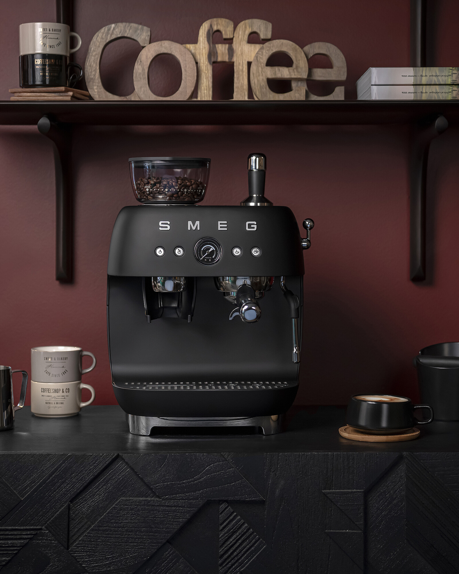 Eine schwarze SMEG-Espressomaschine mit Kaffeebohnen in einem Glasbehälter. Kaffeetassen und ein hölzernes "Coffee"-Schild sind sichtbar.