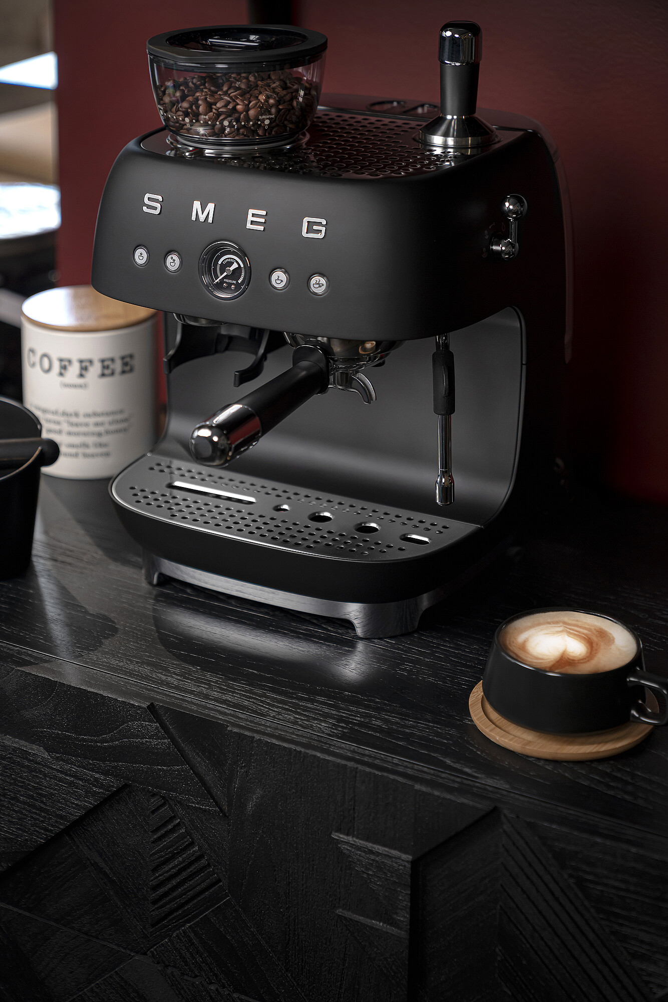 Eine schwarze SMEG-Espressomaschine mit Kaffeebohnen, eine Kaffeetasse mit Latte Art und ein Kaffeebehälter auf Holzoberfläche.