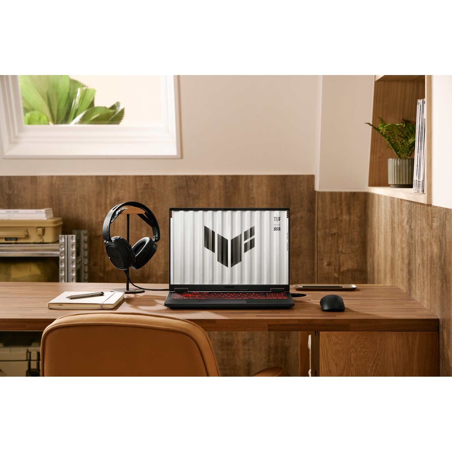 Laptop su scrivania in legno con cuffie, mouse e taccuino. Finestra e pianta sullo sfondo. Sedia davanti.