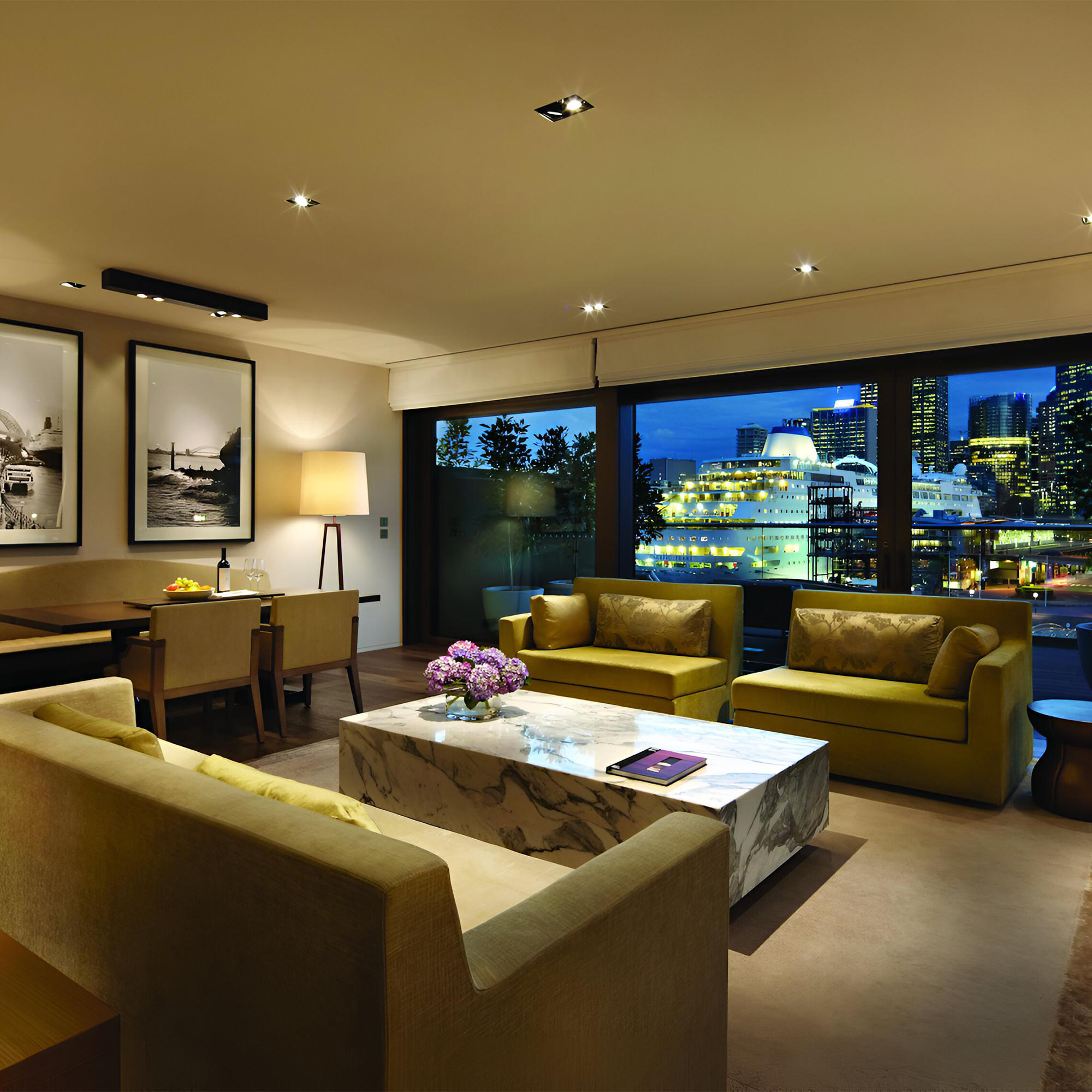 Wohnzimmer mit Stadtblick bei Nacht. Gelbe Sofas, Marmortisch, Kunst und eine Lampe. Schiffe und Gebäude draußen sichtbar.