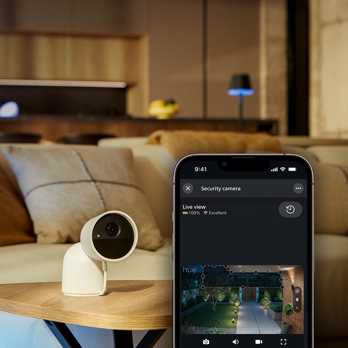 Eine weiße Überwachungskamera steht auf einem Tisch. Ein Telefon zeigt eine Live-Ansicht eines Gartens. Sofa und Lampe sind zu sehen.