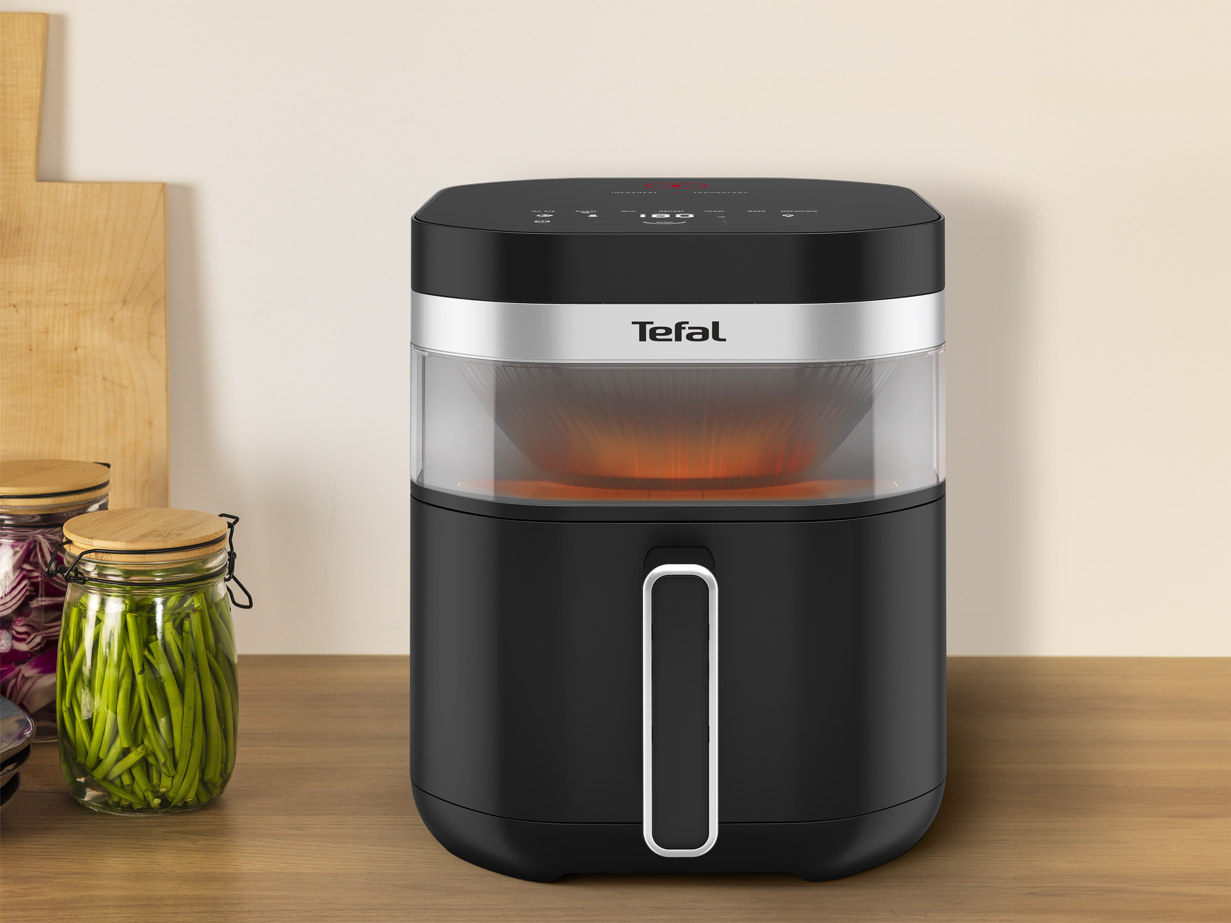 Fekete Tefal légkeveréses sütő fa felületen, élelmiszeres üvegekkel és vágódeszkával a háttérben.