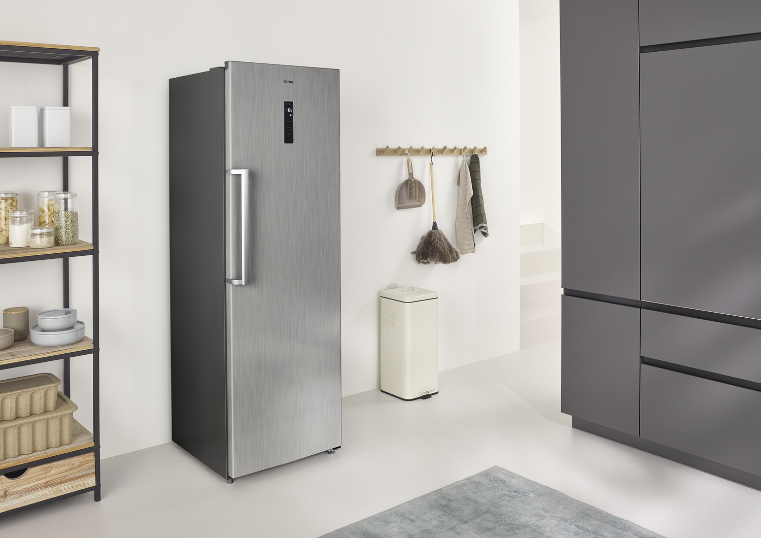 Un refrigerador de acero inoxidable está en una cocina. Un estante de madera con frascos, un recogedor, una escoba y un bote de basura son visibles.