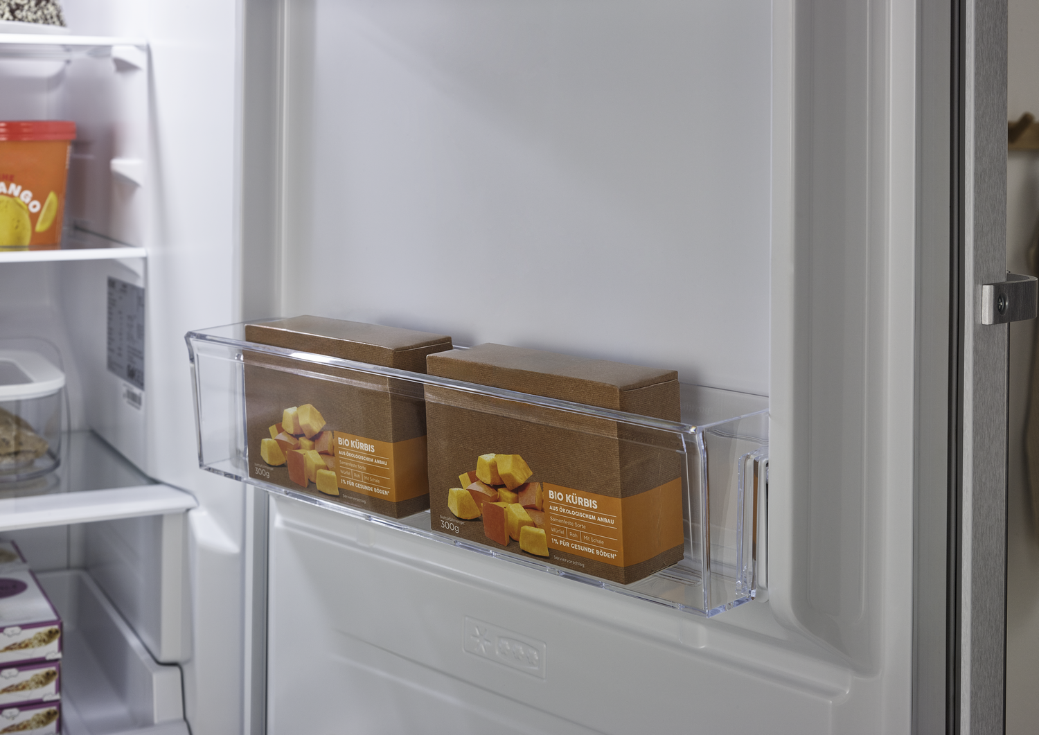 Dentro de un refrigerador, dos cajas de 'BIO KÜRBIS' están en un recipiente transparente. Se ven jugo de mango y otros alimentos.