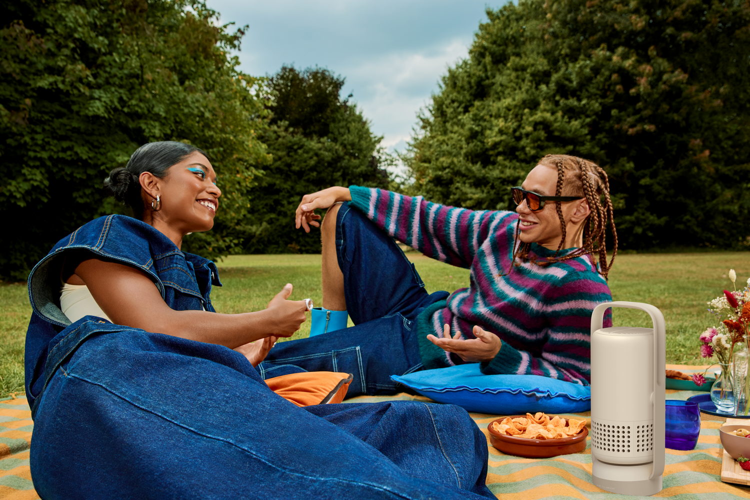 Dos personas en un picnic, sonriendo. Uno lleva mezclilla, el otro un suéter a rayas. Hay aperitivos, flores y un altavoz.
