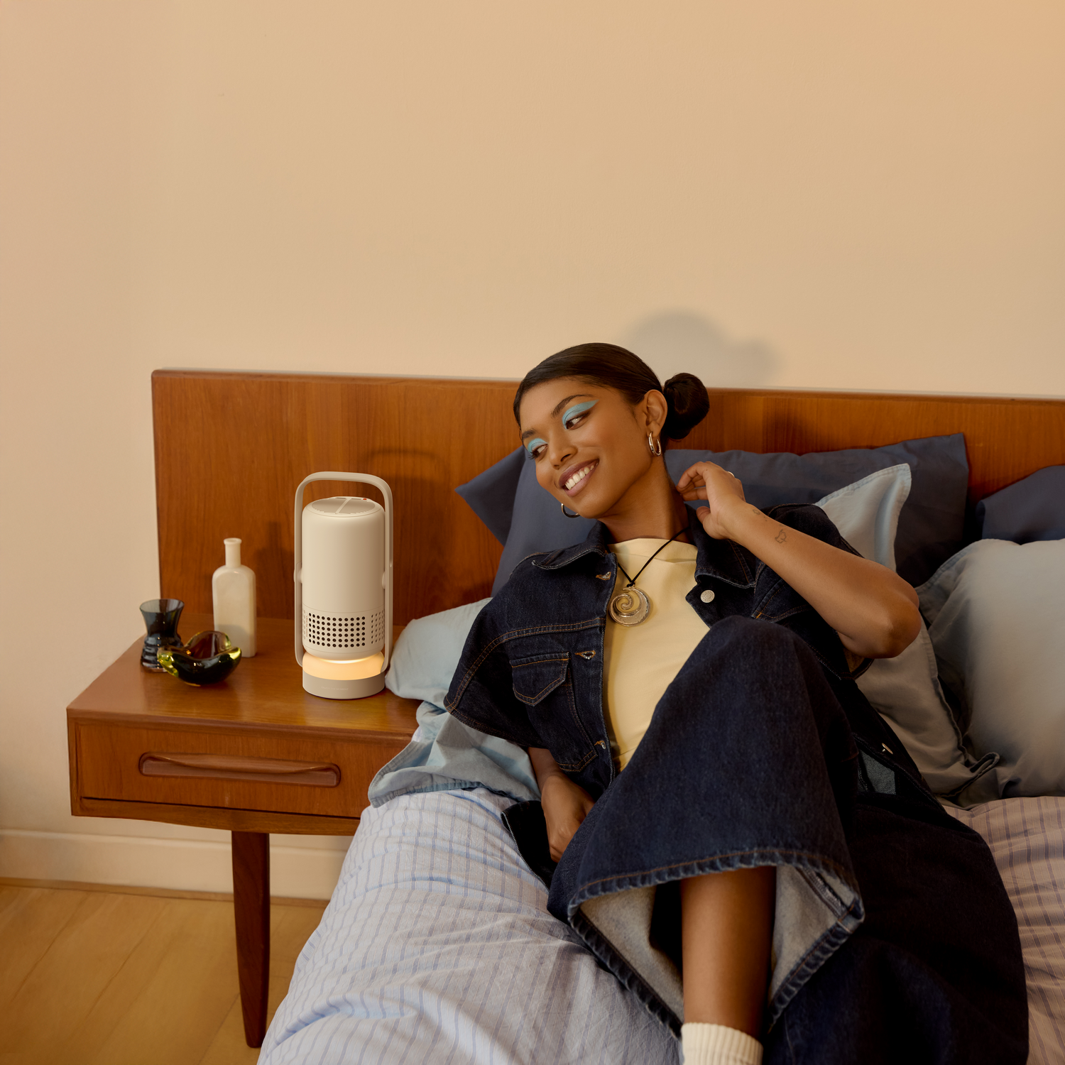Mujer en la cama sonríe, chaqueta vaquera. Purificador de aire blanco en la mesita de noche. Ropa de cama azul y rayada, cabecero de madera.