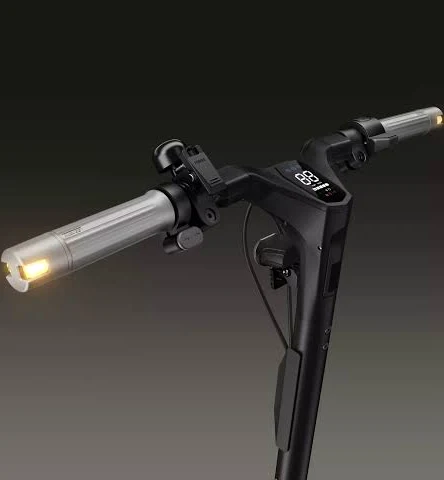 Primer plano del manillar de un patinete eléctrico. El patinete es negro con asas plateadas y una pantalla iluminada. El fondo es oscuro.