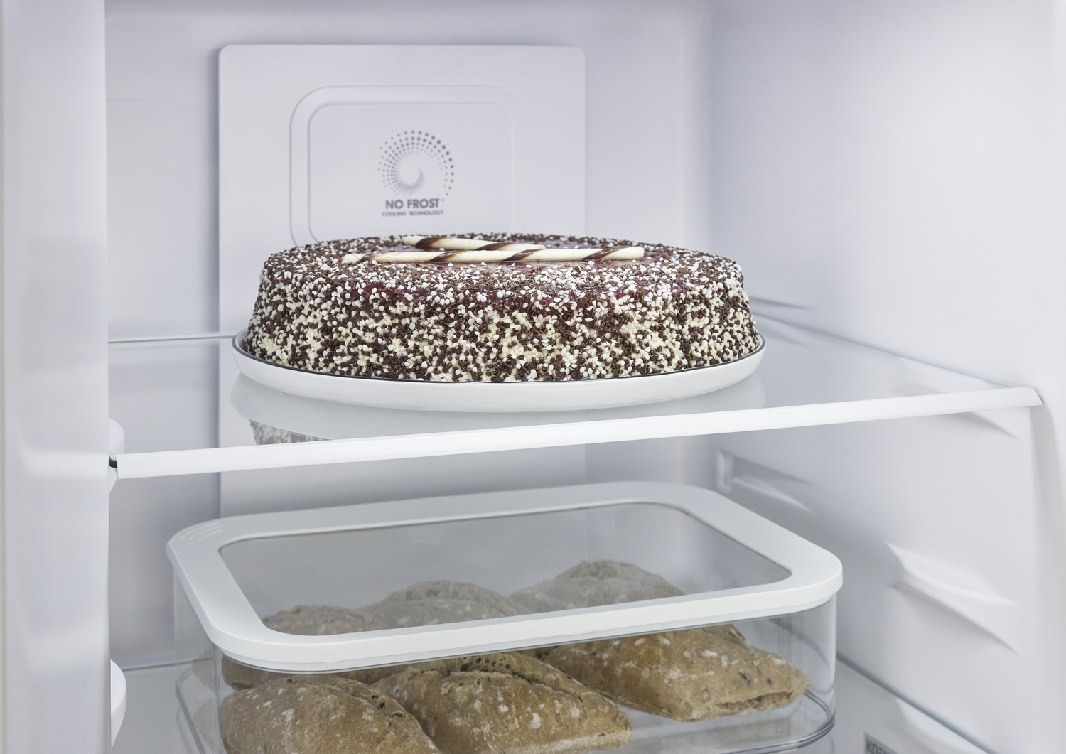 Dentro de un refrigerador, un pastel en un plato y panecillos en un recipiente. El pastel es redondo y decorado.