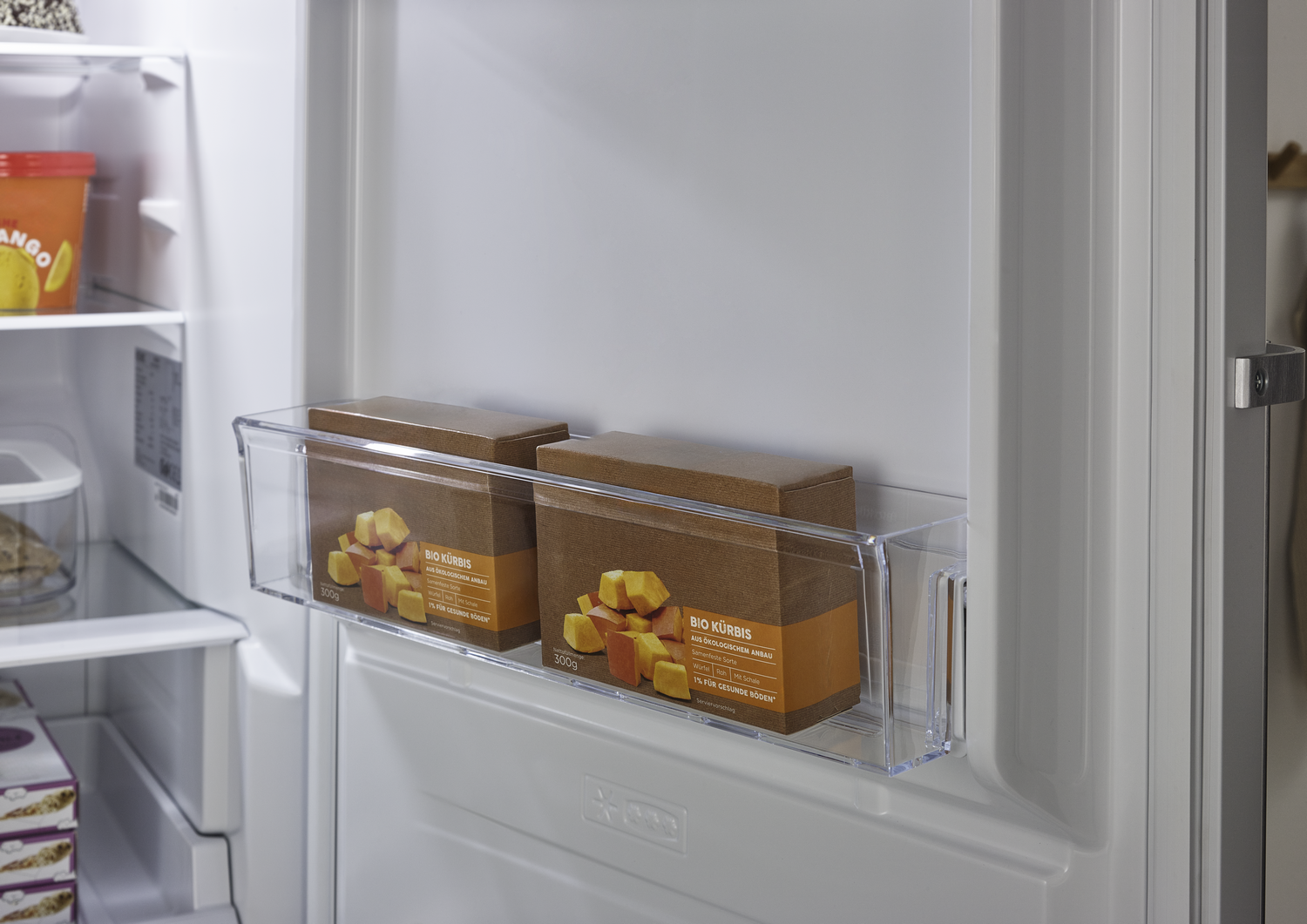 Dentro de una nevera, hay dos cajas de calabaza orgánica en un recipiente transparente. También se ve jugo de mango naranja y otros alimentos.