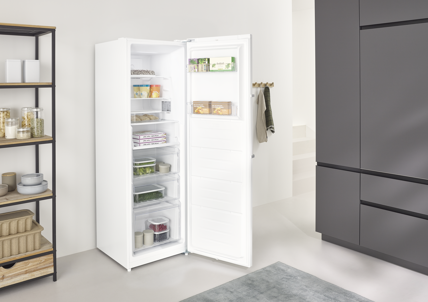 Refrigerador blanco abierto con comida dentro. Se ven estantes, cajones y varios productos. Un estante y un armario gris al fondo.