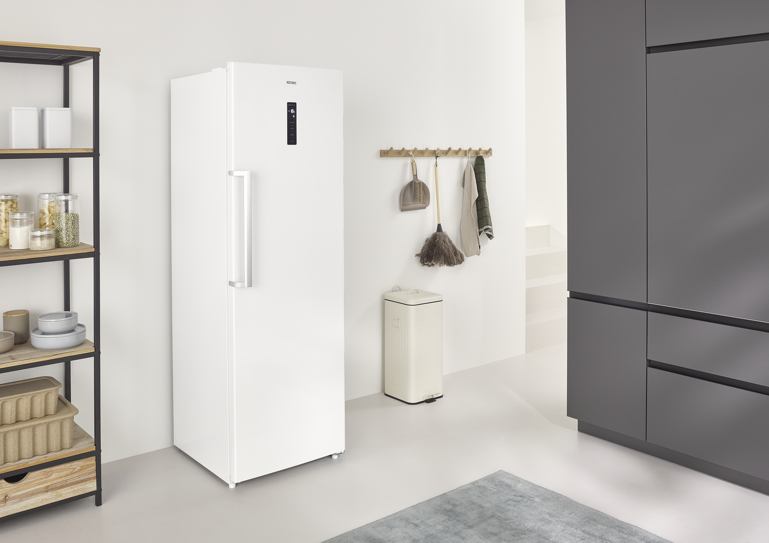 Un refrigerador blanco junto a una estantería con objetos. Una escoba, recogedor y toalla cuelgan de la pared. Una alfombra gris está en el suelo.