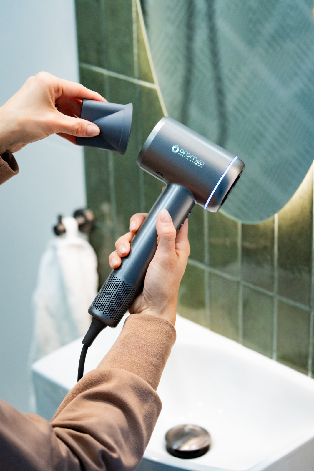 Ręce trzymające szarą suszarkę do włosów, zakładające dyszę. Łazienka z umywalką i lustrem w tle.