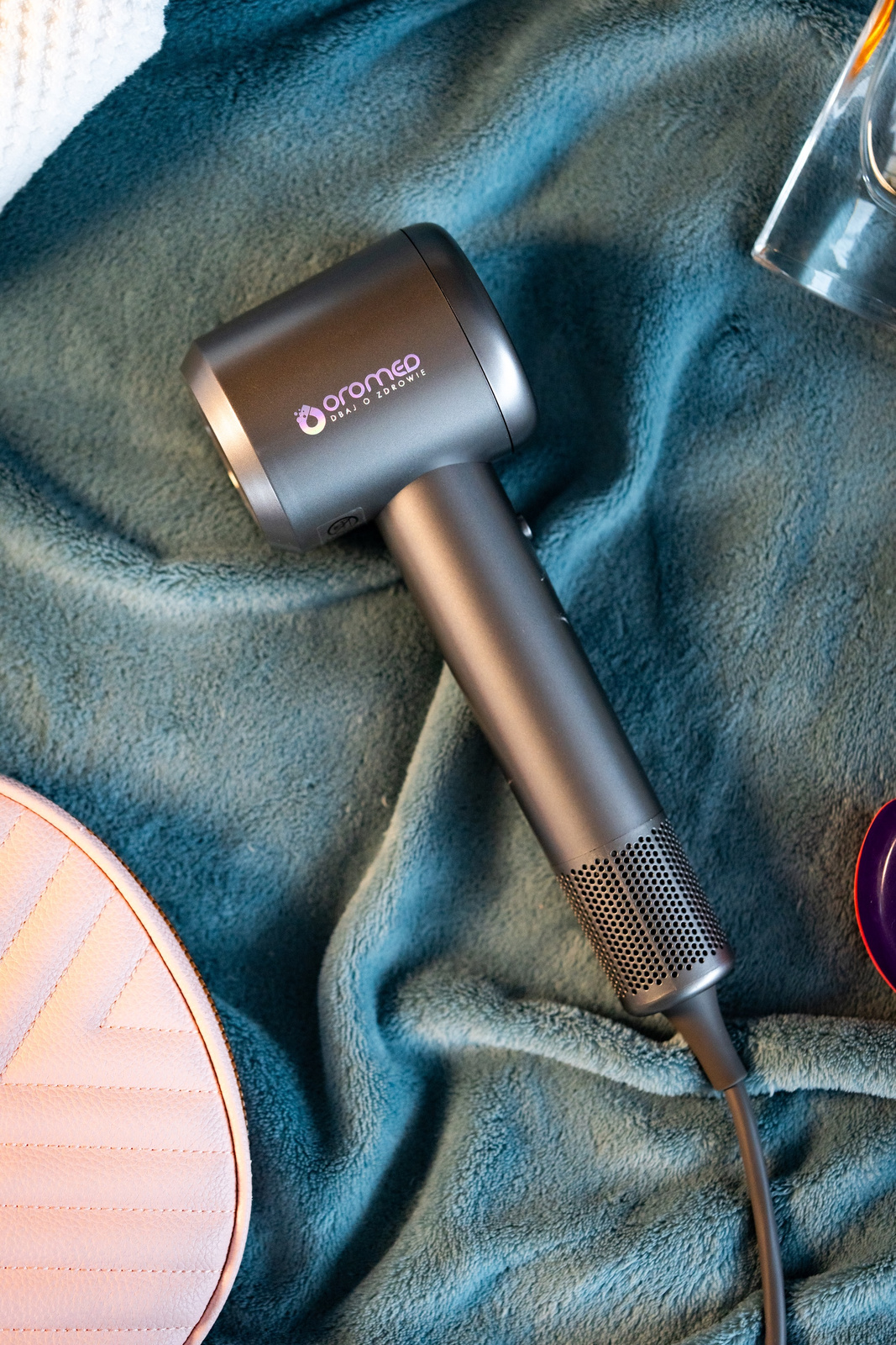 Szara suszarka do włosów z marką 'aromeo' leży na turkusowym kocu. Widoczna jest również różowa torba i szklanka.
