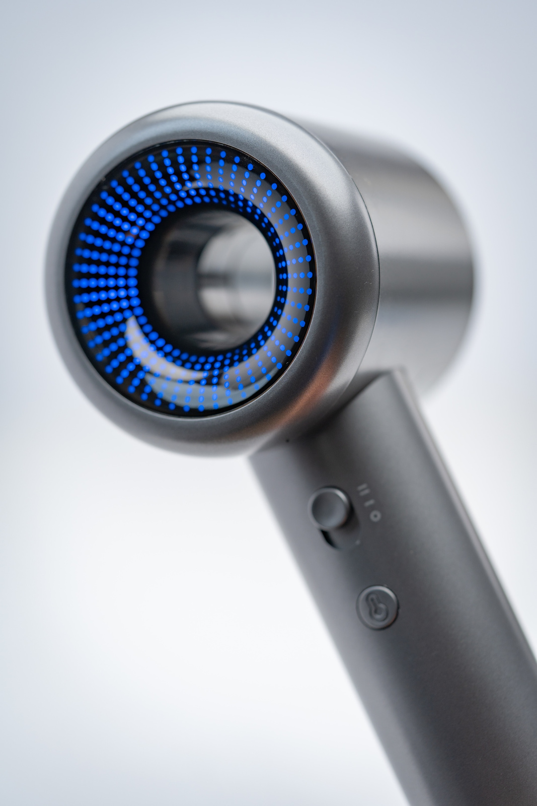 Zbliżenie szarej suszarki do włosów. Dysza ma niebieskie kropki. Uchwyt ma przyciski i przycisk zasilania. Białe tło.