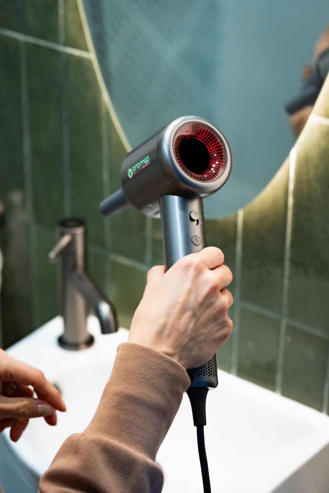 Osoba trzyma srebrno-czarną suszarkę do włosów w łazience. Suszarka jest skierowana w stronę lustra. Zlew jest w tle.